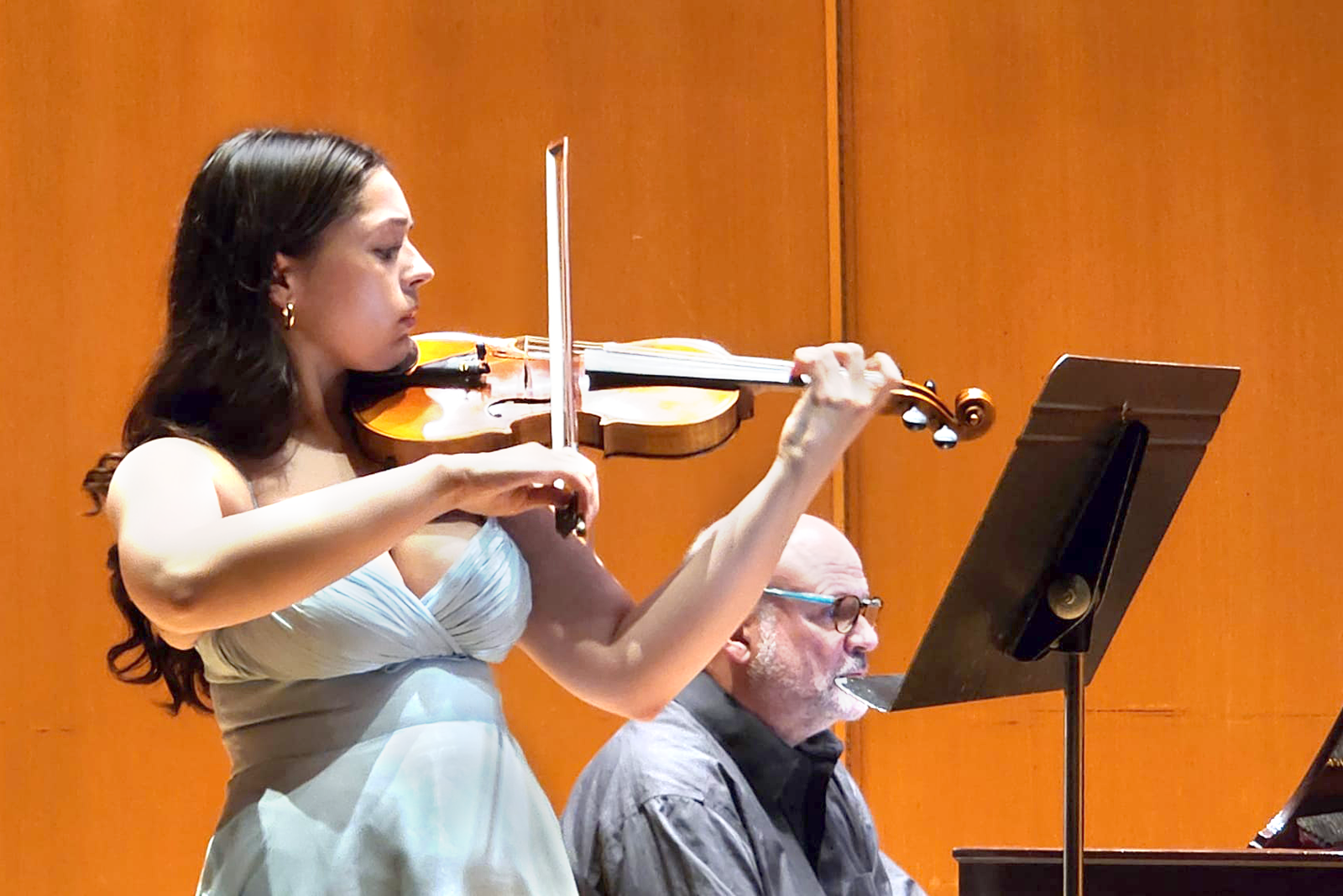 a young woman with long, dark hair playing violin on a stage in front of a man sitting at a piano