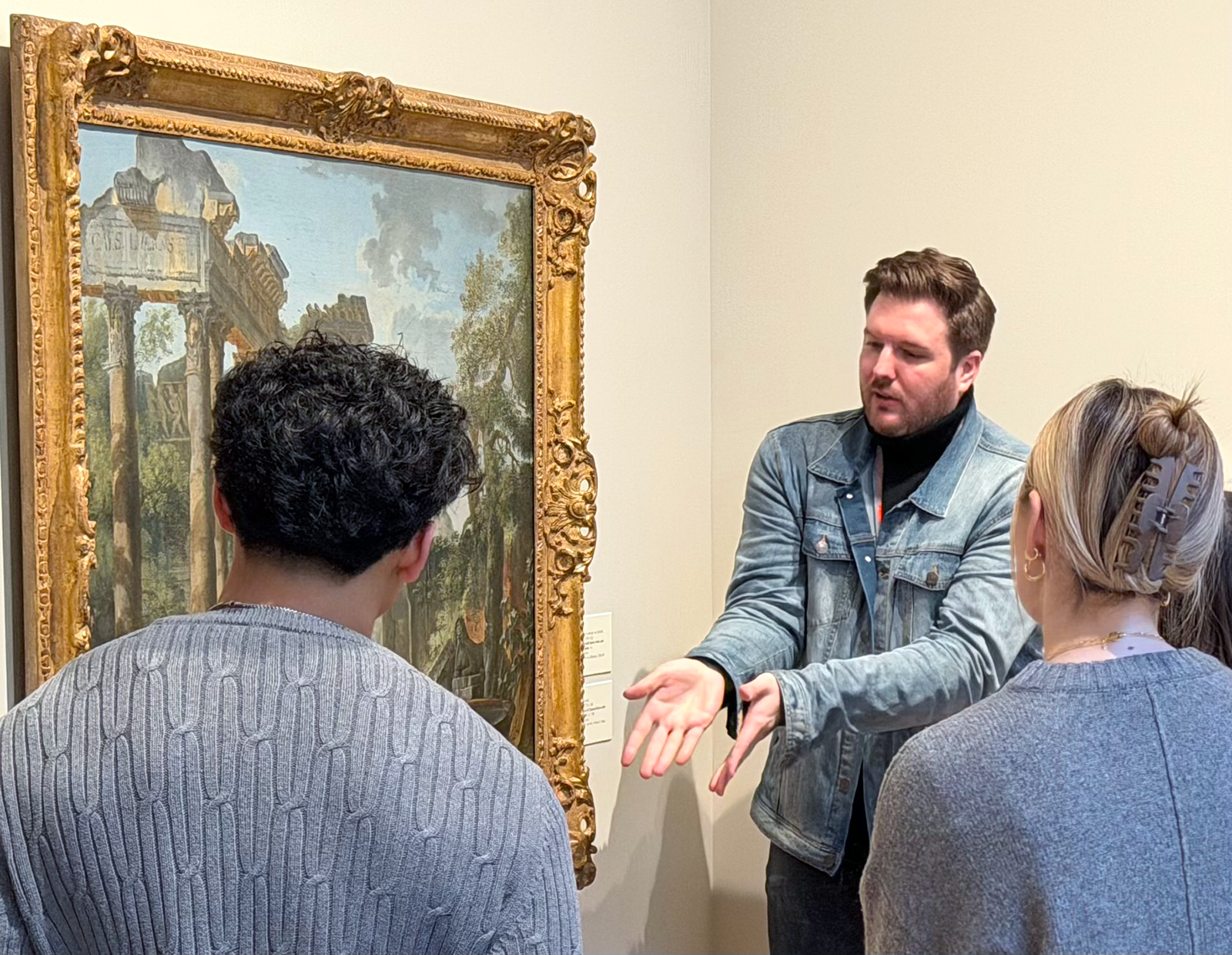 a man in a denim jacket gestures toward a large, ornately framed painting, with the backs of onlooking students' heads in the foreground